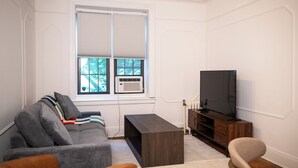 Una sala de estar con un sofá gris, una mesa de centro de madera, un televisor sobre un mueble de madera y una ventana con persianas.