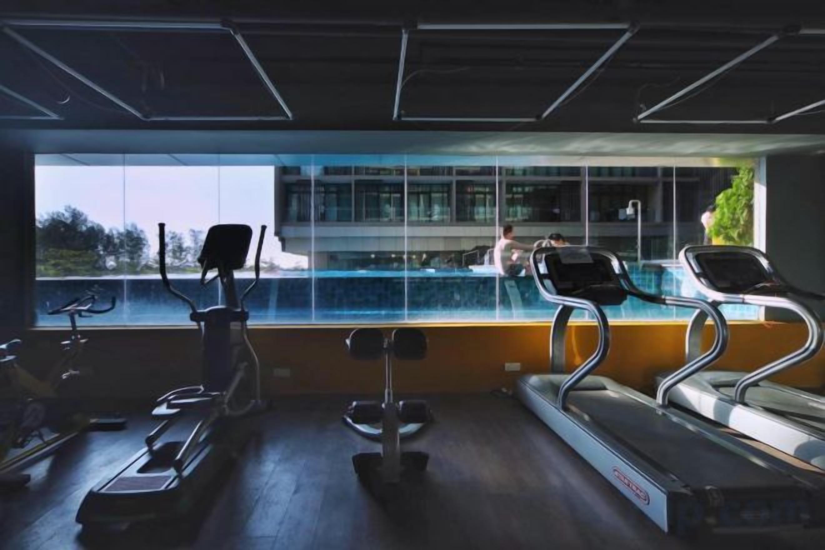 A gym with treadmills, a rowing machine, and a bench press. There is a large window with a view of a building and greenery outside.