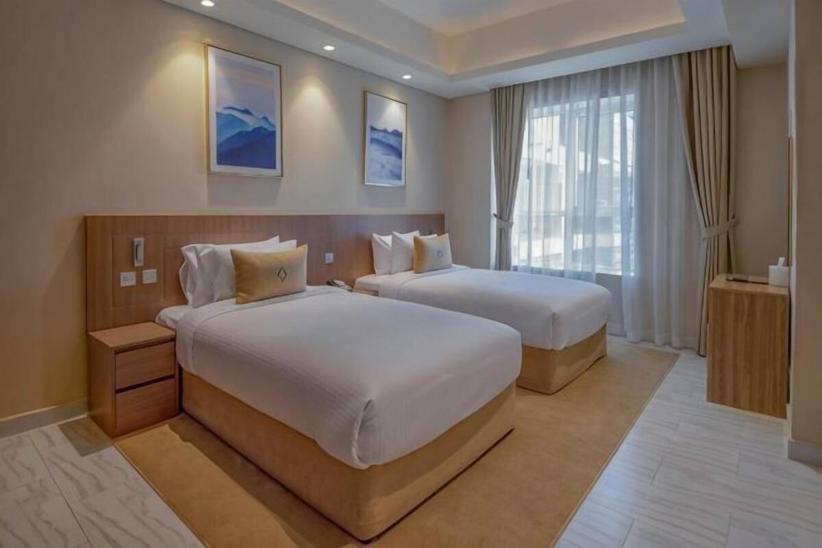 Habitación de hotel con dos camas, una mesita de noche, una pequeña mesa y un ventanal con cortinas.