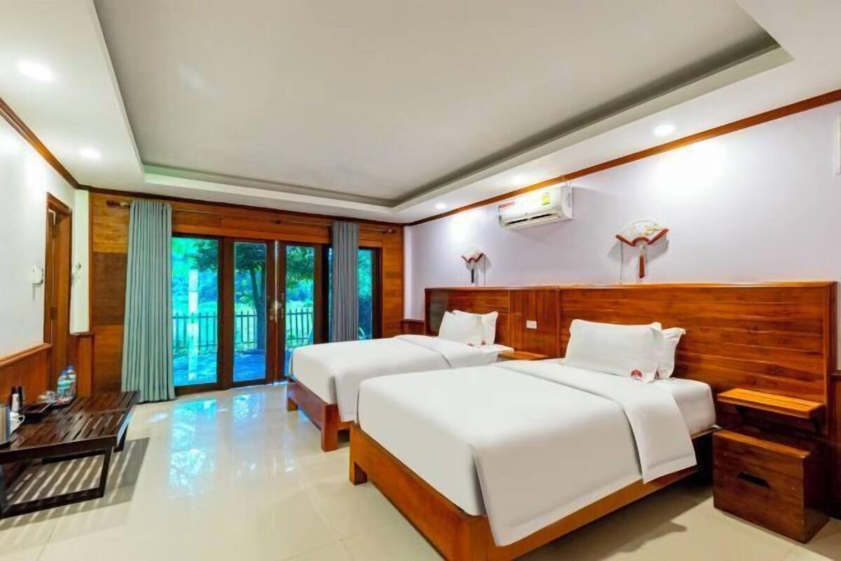 Une chambre d’hôtel avec deux lits, une tête de lit en bois, un bureau et une vue sur la verdure à travers les portes vitrées.