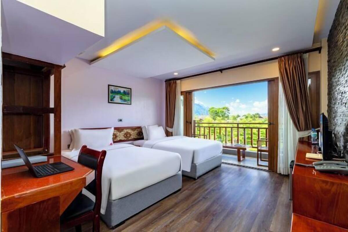 Une chambre d’hôtel avec deux lits, un bureau avec un ordinateur portable, une télévision et un balcon donnant sur un espace verdoyant.