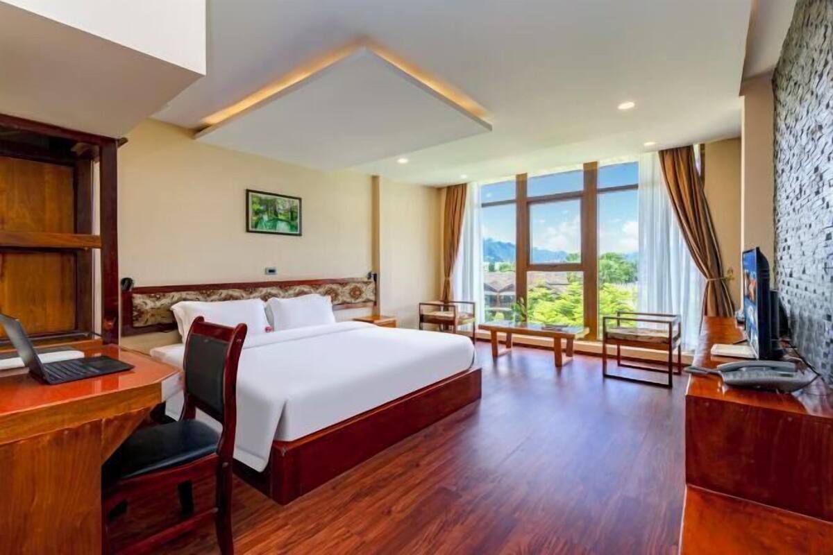Une chambre d’hôtel avec un grand lit, un bureau avec un ordinateur portable, une chaise, une petite table et une vue sur l’extérieur grâce à de grandes fenêtres.