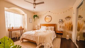 Un dormitorio con cama, mesitas de noche, un escritorio, una silla, ventilador de techo, ventana con cortinas, un espejo y plantas decorativas.