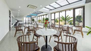 A spacious indoor area with multiple round tables and chairs, large windows, and a staircase leading to an upper level.
