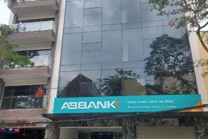 Egy épület, amelyen az „ABBANK” feliratú tábla látható, és amelynek üveg homlokzatán a fák képe tükröződik.