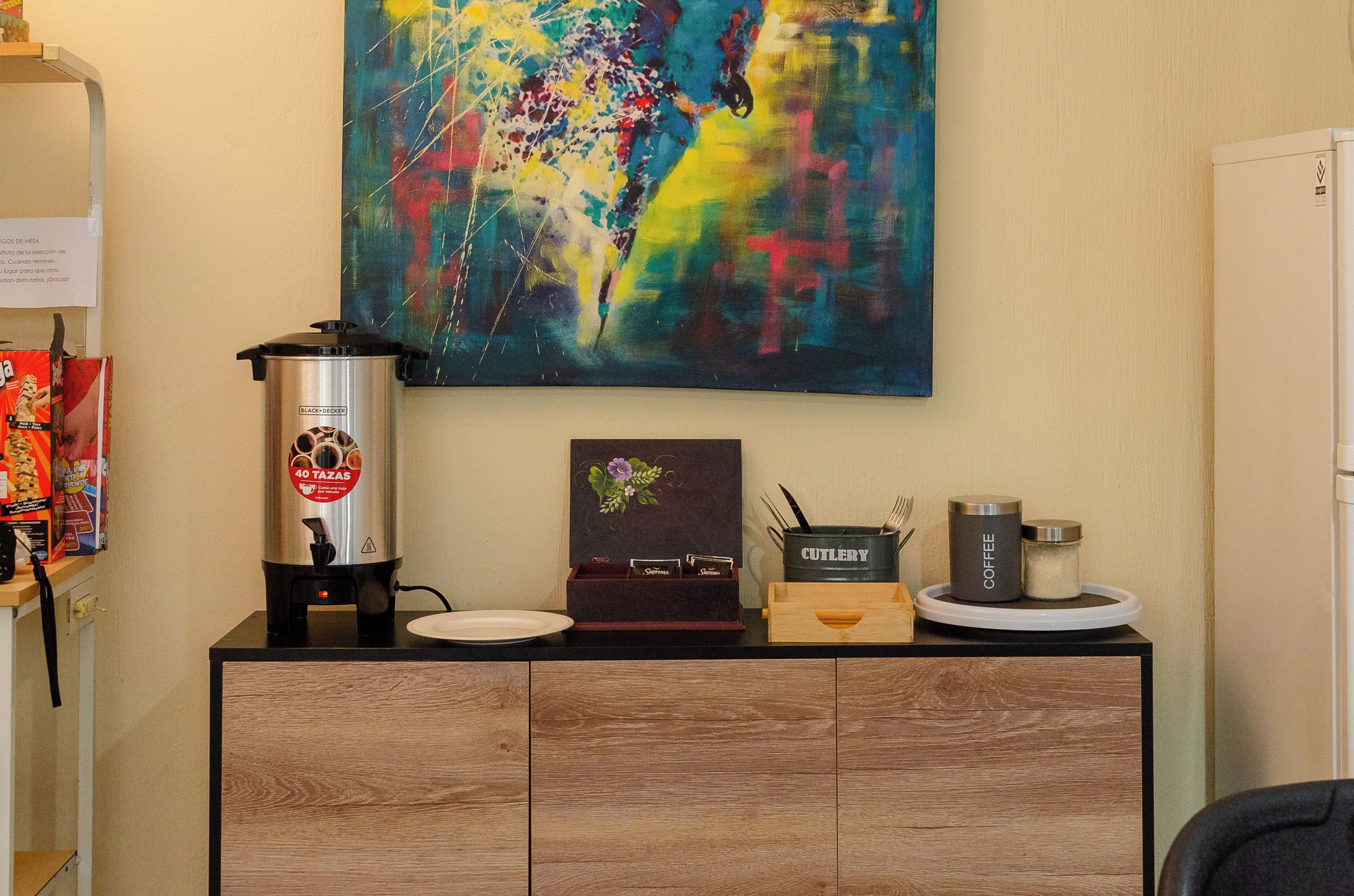 A kitchenette with a coffee maker, a plate, a cutlery holder, and a coffee jar on a wooden cabinet.