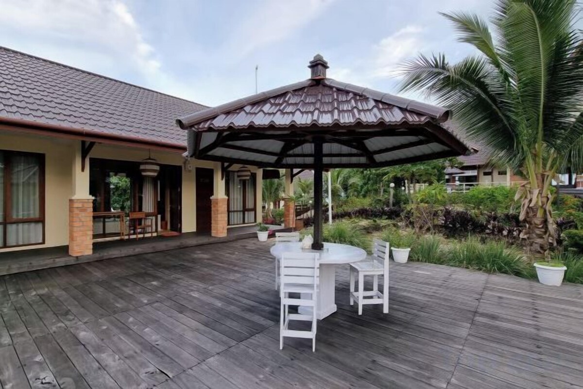 A covered outdoor area with a wooden deck, a dining table with chairs, and a palm tree.