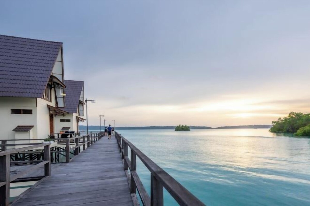 A wooden pier leading to overwater bungalows with a calm sea and sunset in the background.