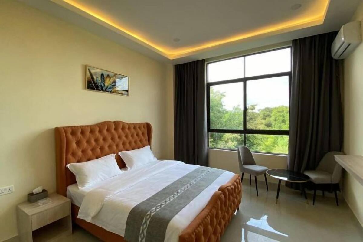 A hotel room with a large bed, a desk, two chairs, a window with curtains, and a painting on the wall.