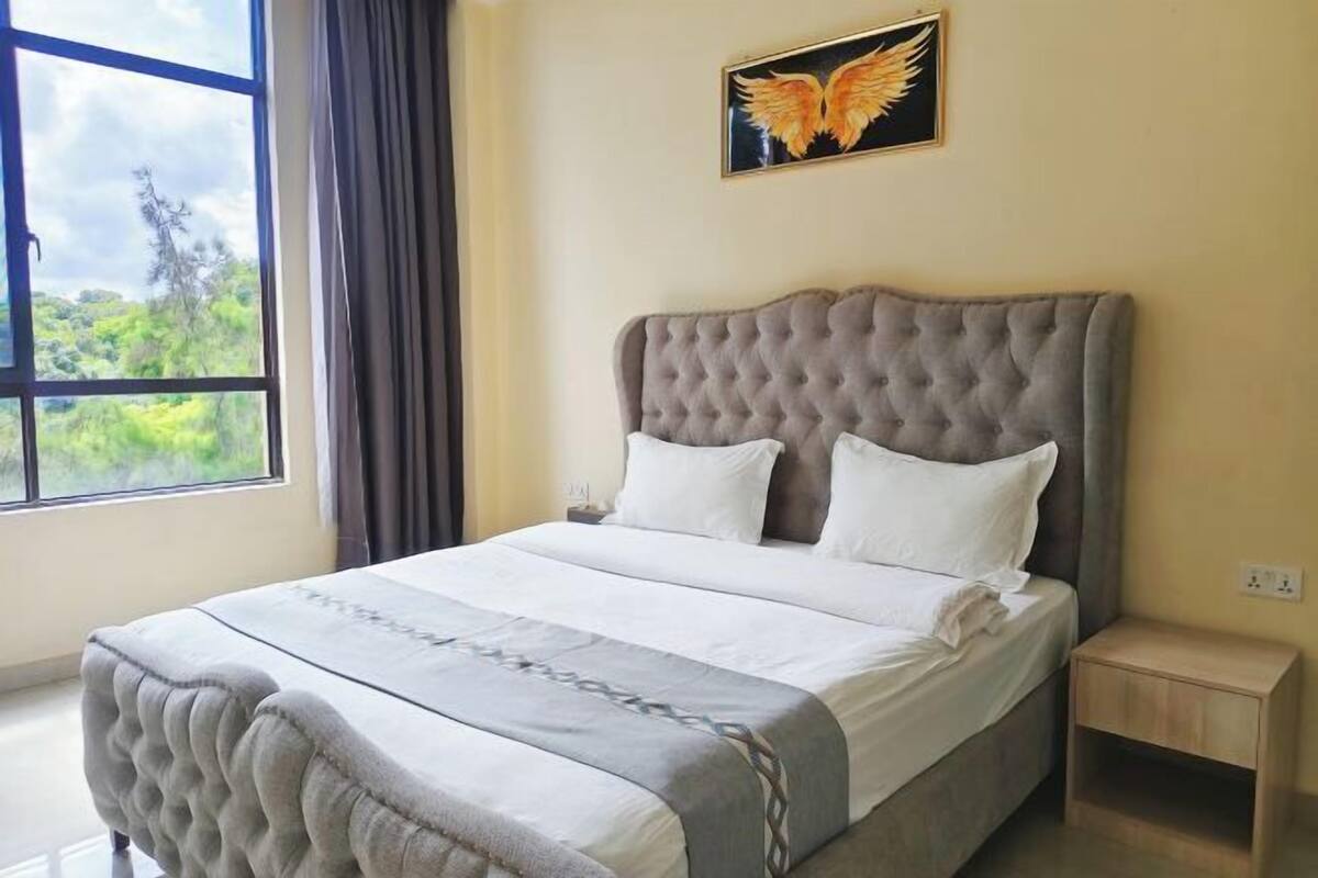 A neatly made bed with a grey headboard and white bedding, a small bedside table, and a window with a view of greenery.