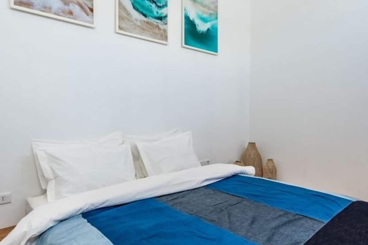 Una cama con ropa de cama blanca y azul, una mesita de noche de madera y dos cuadros colgados en la pared.
