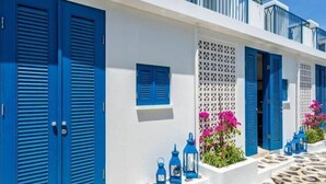 Ein weißes Gebäude mit blauen Fensterläden und -türen, ein Balkon mit Geländer sowie ein mit Steinplatten gepflasterter Innenhof.