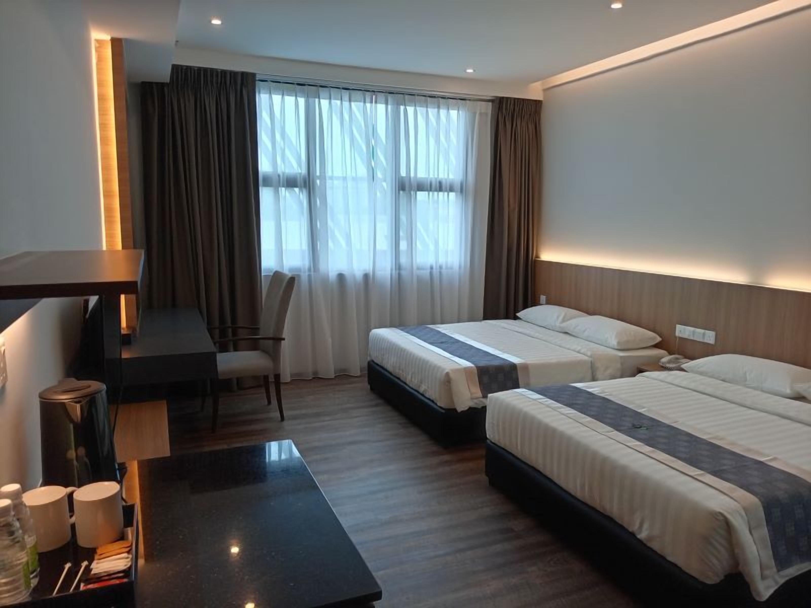 A hotel room with two beds, a desk, a chair, and a table with cups and a bottle.