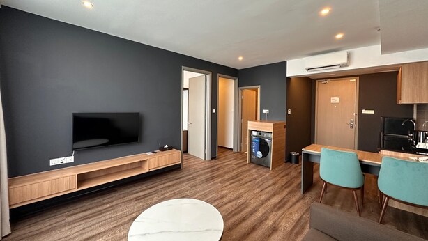 Um apartamento moderno com televisão de tela plana, uma sala de jantar com duas cadeiras, uma cozinha com geladeira preta, e máquina de lavar e secar embutidas.