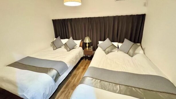 A hotel room with two beds, a bedside table with a lamp, and a large window with curtains. - Osaka Nimi family house direct KIX airport (Osaka)