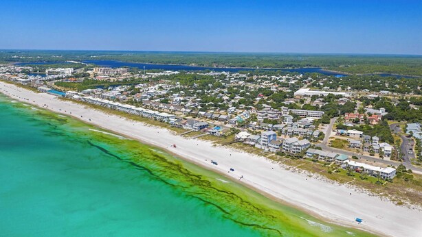 An aerial view of a coastal town with a long beachfront, dense residential area, and a large body of water in the background.
