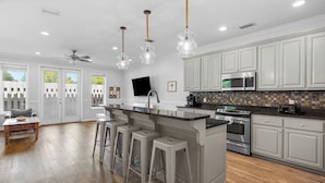 A modern kitchen with white cabinets, a central island with bar stools, a microwave, stove, and a flat-screen TV mounted on the wall.