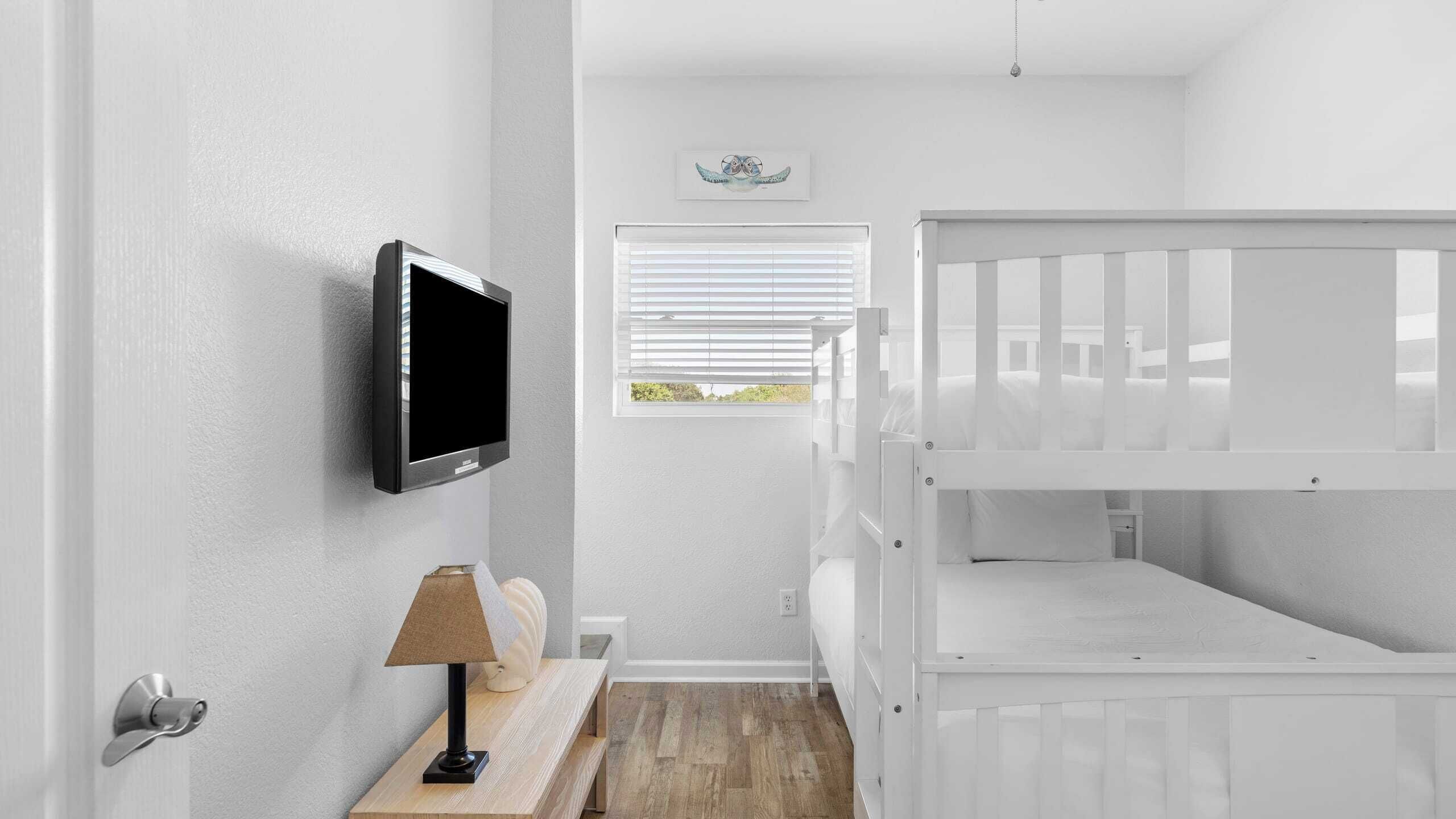 A room with a white crib, a mounted TV, a window with blinds, and a small table with a lamp.
