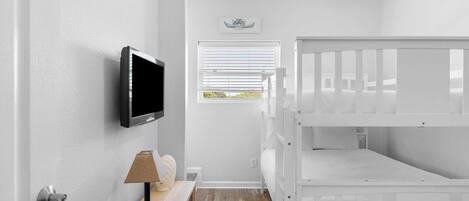 A room with a white crib, a mounted TV, a window with blinds, and a small table with a lamp.