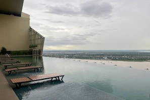 An infinity pool with wooden loungers and a view of a beach and cityscape. - The Peak Service Apt By David (Phnom Penh)