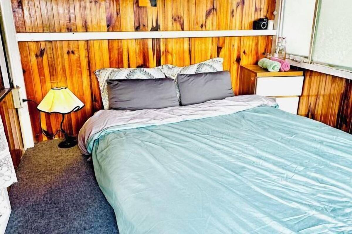 A bedroom with a wooden headboard, a bed with a blue bedspread, two pillows, a bedside table with a lamp, and a window with a view.