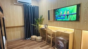 A room with a wooden floor, a desk with a chair, a television mounted on the wall, a potted plant, and a box on the floor. - Ben Thanh Luxury Suite Hotel (Ho Chi Minh City)