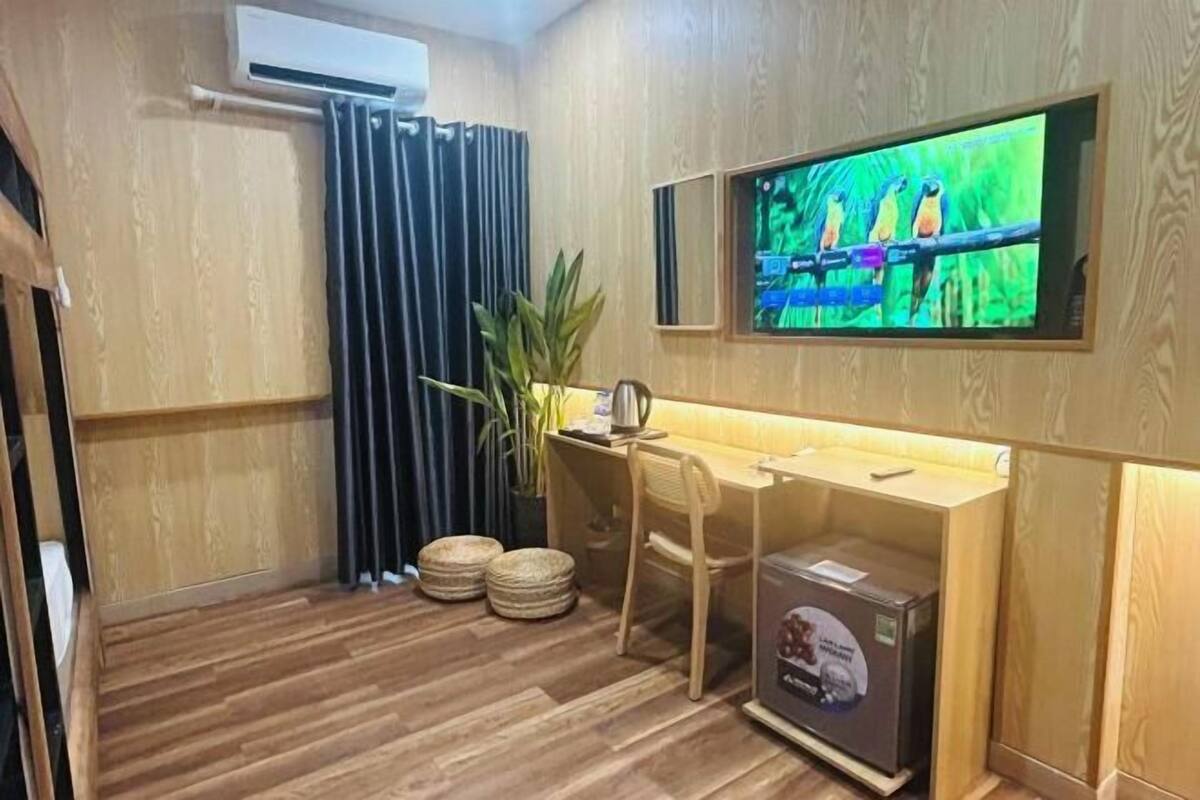 Une pièce avec un plancher en bois, un bureau avec une chaise, un téléviseur fixé au mur, une plante en pot et une boîte posée sur le sol.