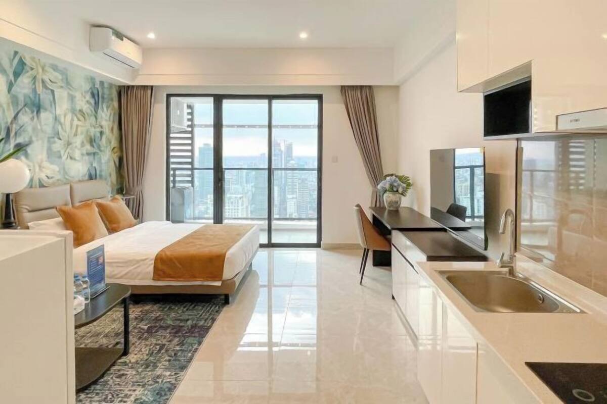 Une chambre d’hôtel moderne équipée d’un lit, d’un bureau, d’une kitchenette et d’une grande fenêtre donnant sur la ville.