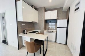 Une cuisine compacte avec un plan de travail blanc, un évier et une cuisinière. On y trouve une chaise et un réfrigérateur.