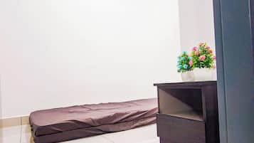 A room with a mattress against a wall, a dark-colored cabinet with a potted plant on top, and a tiled floor.