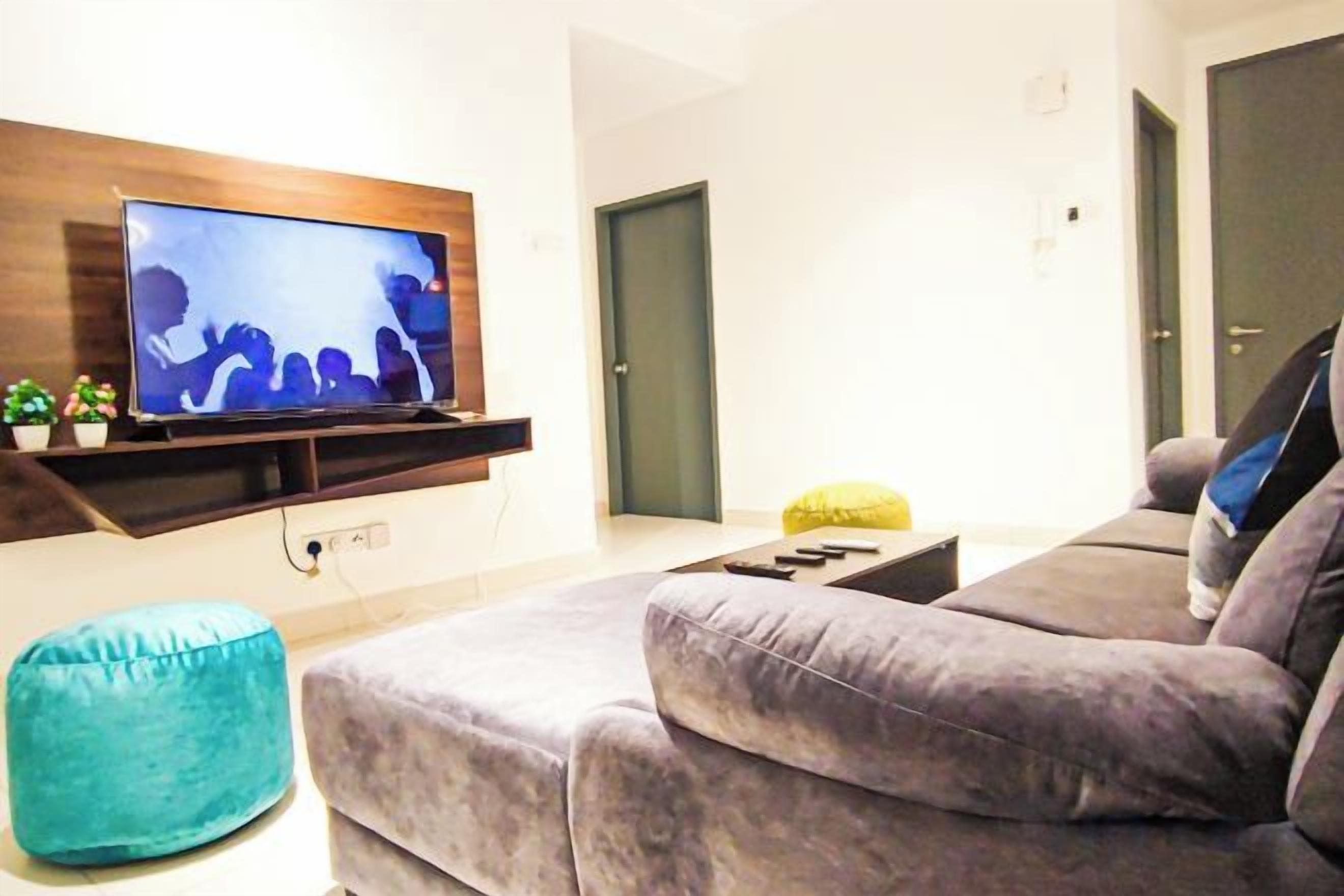 Ett vardagsrum med en platt-TV som är monterad på en hylla i trä, en grå soffa, en turkos sittpuff och ett litet bord med ett gult föremål på.