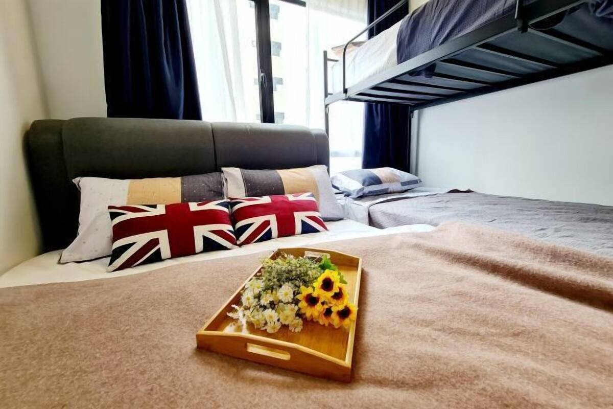 Quarto de hotel com uma cama, mesa de cabeceira, um tabuleiro com flores e uma beliche ao fundo.