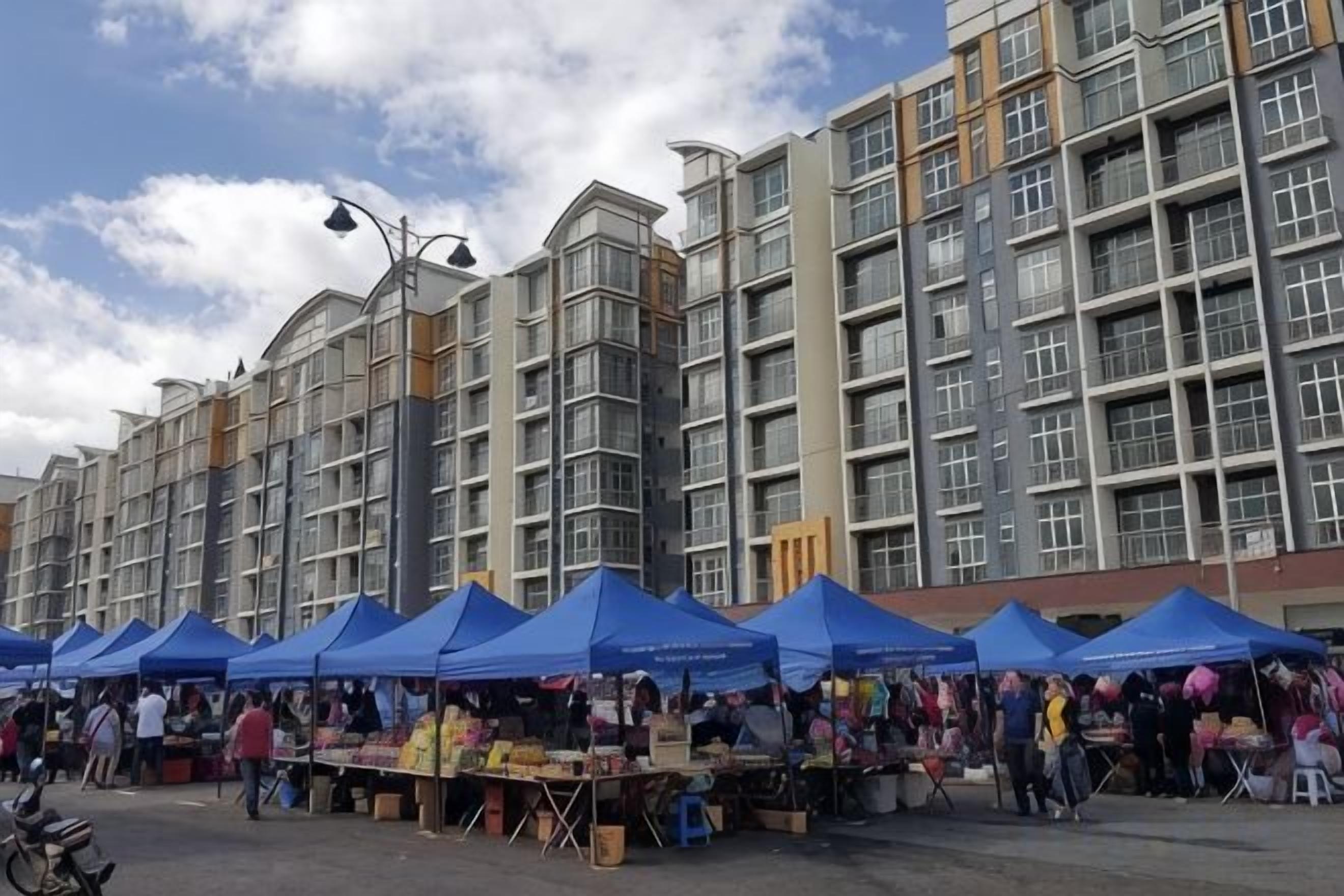 A market with blue tents and a multi-story building in the background.