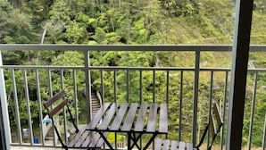 Een balkon met een houten tafel en stoel, omgeven door een metalen hek, met uitzicht op een dicht bos.