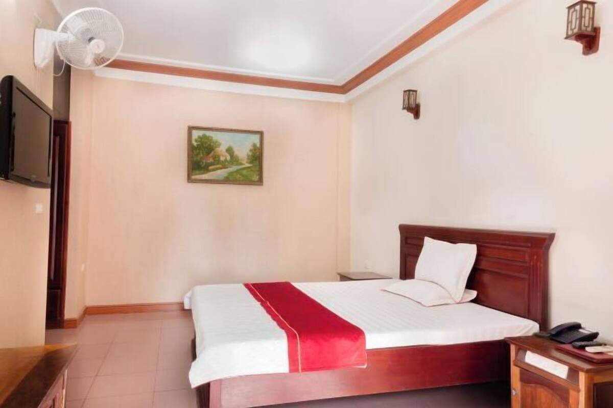 Une chambre d’hôtel avec un lit, un ventilateur, un tableau au mur et une table de chevet avec un téléphone.