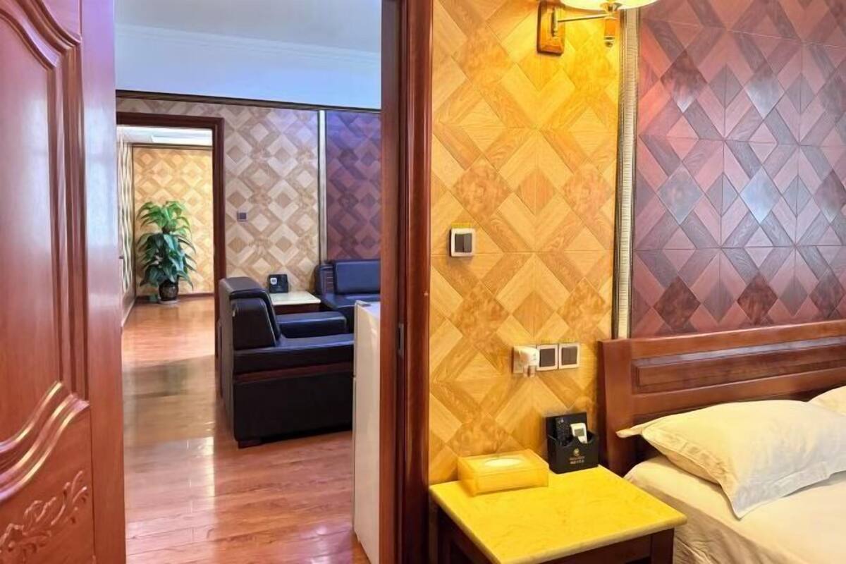 A hotel room with a wooden headboard, a bedside table, a bed, a seating area with a sofa, and a wall with a geometric pattern.