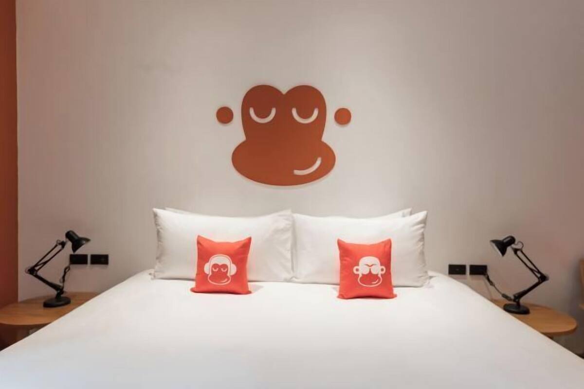 Une chambre d’hôtel avec un lit, deux oreillers ornés de motifs de singes, une décoration murale représentant un visage de singe et deux lampes de chevet.