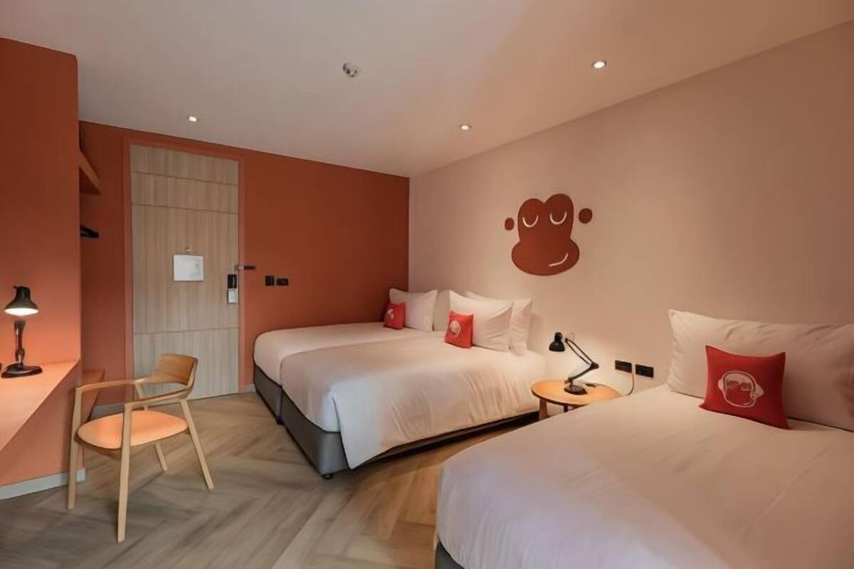 Une chambre d’hôtel avec deux lits, un bureau et une chaise. Il y a un élément décoratif sur le mur et une porte avec un rideau.