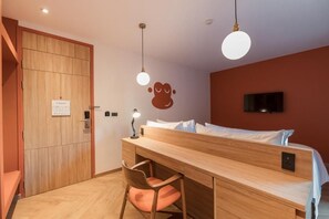 A room with a wooden desk, a chair, a bed, a wall-mounted TV, and a door with a sign.