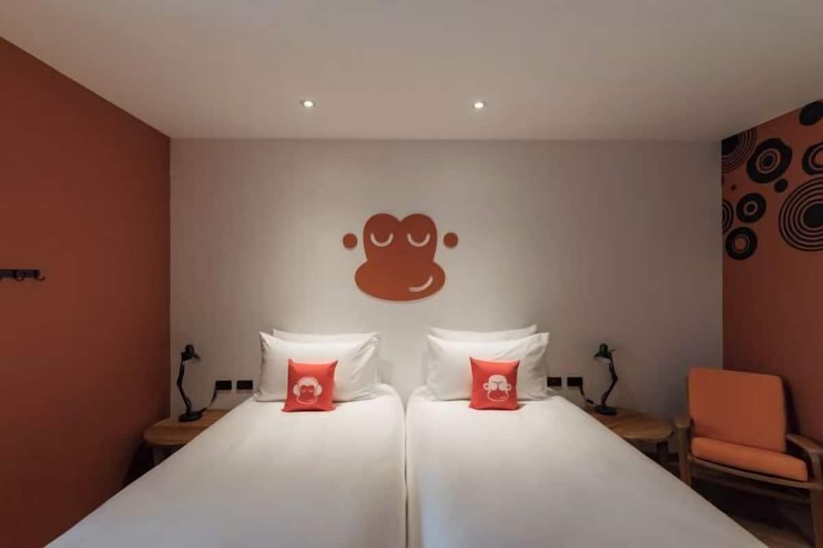 Une chambre d’hôtel avec deux lits, chacun agrémenté de draps blancs et d’un oreiller rouge. Au-dessus des lits, on trouve une œuvre d’art accrochée au mur, une petite table avec une lampe et une chaise orange.