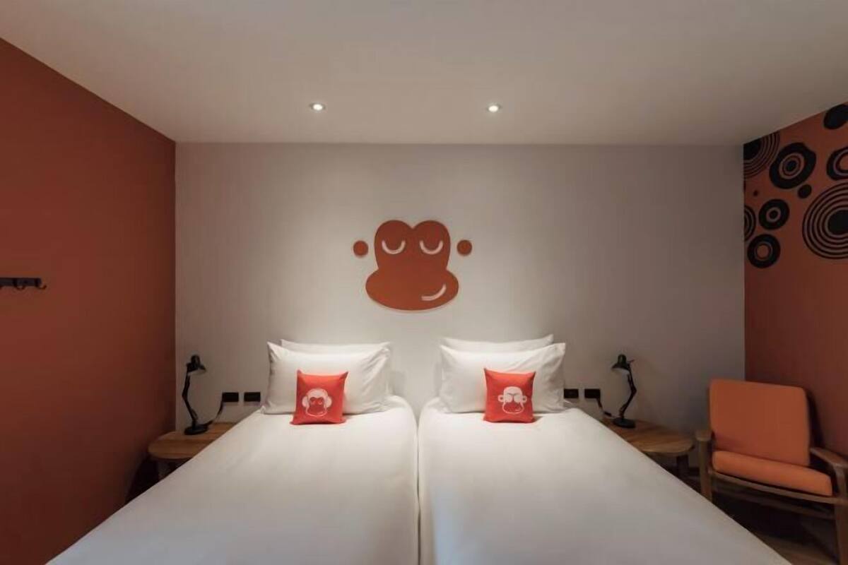 Ein Hotelzimmer mit zwei Betten, jeweils mit weißer Bettwäsche und einem roten Kissen. Darüber befindet sich ein Wandbild, ein kleiner Tisch mit einer Lampe und ein orangefarbener Sessel.