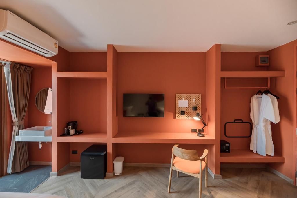 A room with a TV mounted on the wall, a desk with a chair, a small shelf with a lamp, and a coat rack with a robe.