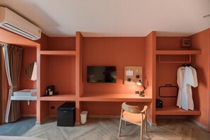 A room with a TV mounted on the wall, a desk with a chair, a small shelf with a lamp, and a coat rack with a robe. - BLU Monkey Hub & Hotel Bangsaen (Chonburi)