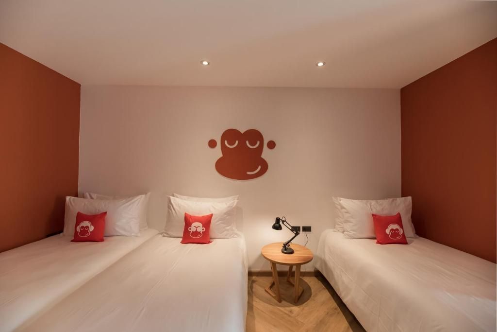 A hotel room with two beds, each with white bedding and red pillows, a small round table with a lamp, and a wall-mounted logo.