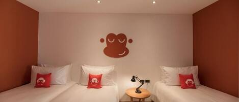Habitació d'hotel amb dos llits, cadascun amb roba de llit blanca i almoades vermelles, una taula rodona petita amb una làmpada i un logotip fixat a la paret.