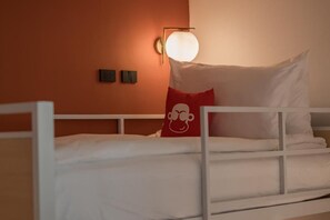 A single bed with white bedding, a red pillow with a cartoon character, and a wall-mounted light fixture. - BLU Monkey Hub & Hotel Bangsaen (Chonburi)