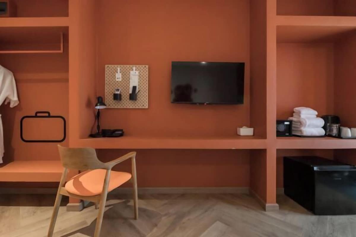 Una camera d'albergo con una sedia di legno, una TV, una bacheca montata a parete e uno scaffale con asciugamani e articoli da toilette.