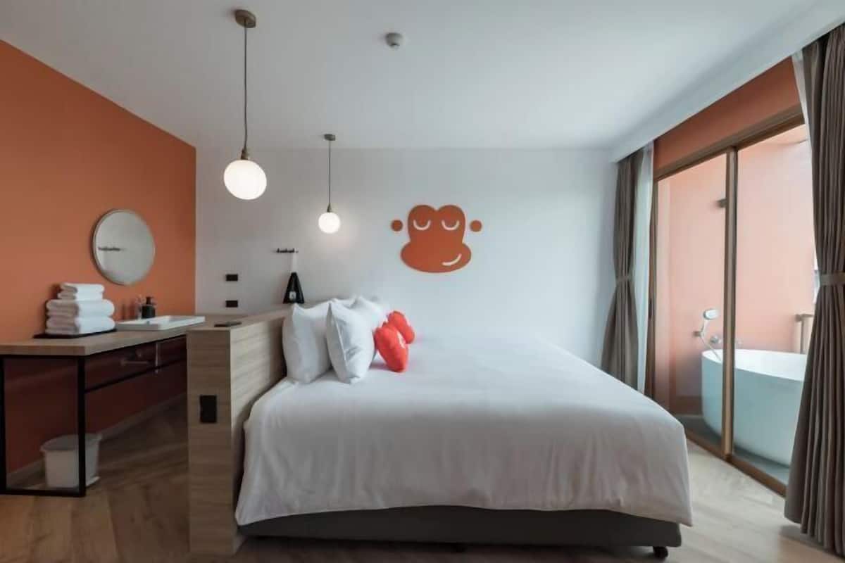 Une chambre d’hôtel moderne avec un grand lit, une table de chevet avec un miroir, une horloge ronde et un luminaire fixé au mur.