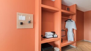 Une chambre d’hôtel avec un mur de couleur pêche, un panneau perforé où sont accrochés divers objets, un réfrigérateur noir, un peignoir blanc et un porte-vêtements suspendu.