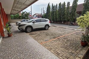 Un espace de stationnement où se trouvent une voiture blanche et une moto, le tout entouré de plantes en pot et situé devant l’entrée d’un bâtiment.